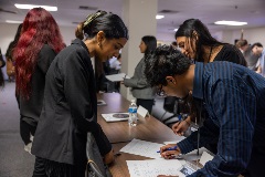 Ayoungwomaninablackblazerandayoungmaninglassesleanoveratablereviewingdocumentstogetherasotherparticipantstalknearby