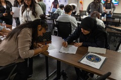 Twowomensitandleanoveratablefillingoutformsonewearingabeigeshirtandtheotherablackheadscarfduringayouthleadershipevent