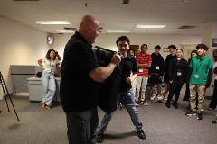 Abaldinstructorholdsapaddedshieldwhileastudentpracticesstrikingtechniquesduringaphysicaltrainingexerciseasotherstudentslookon